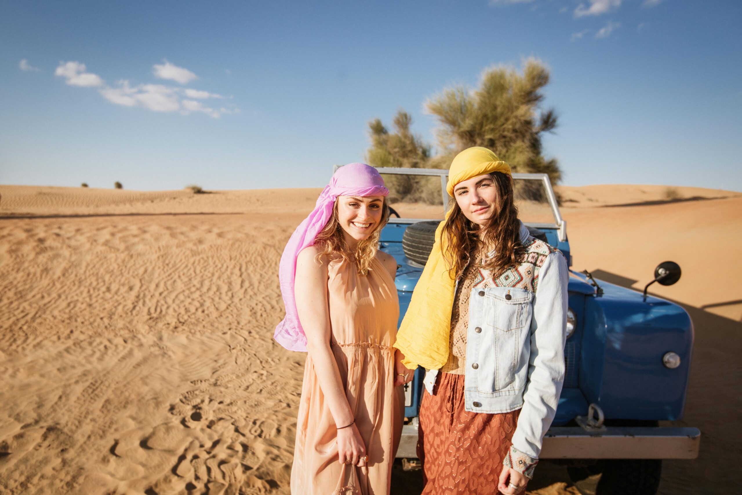 Two women smiling in the Dubai desert with a vintage Land Rover hinting at adventure and travel.
